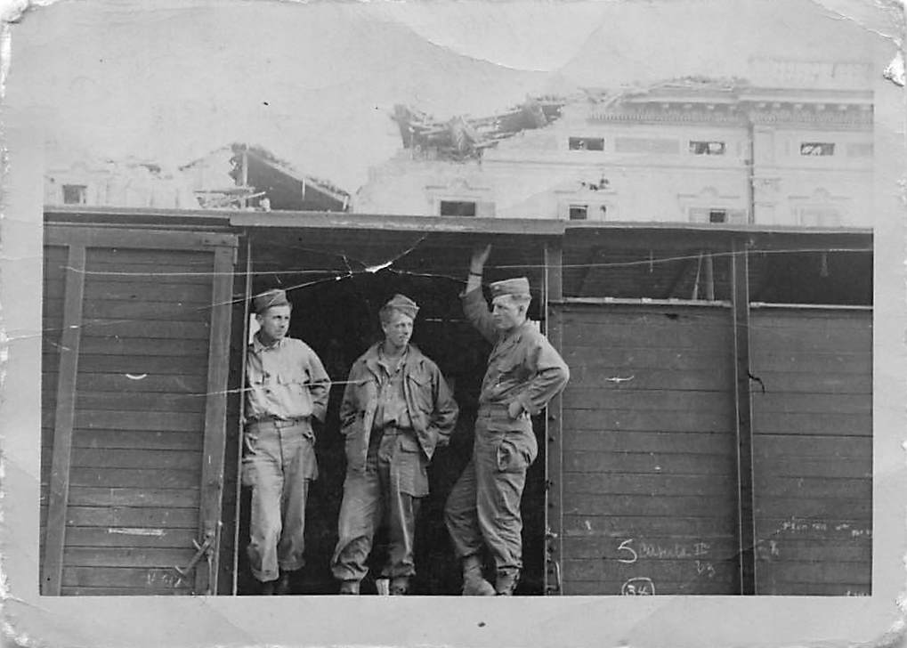 Soldiers in railroad boxcar, bombed city behind